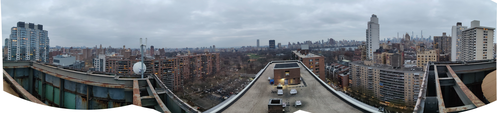 Wide rooftop panorama from a NYC Mesh site looking out across Manhattan: pre-war apartment blocks, gravel rooftops with service bulkheads, Midtown towers in the distant haze, an overcast late-winter sky.