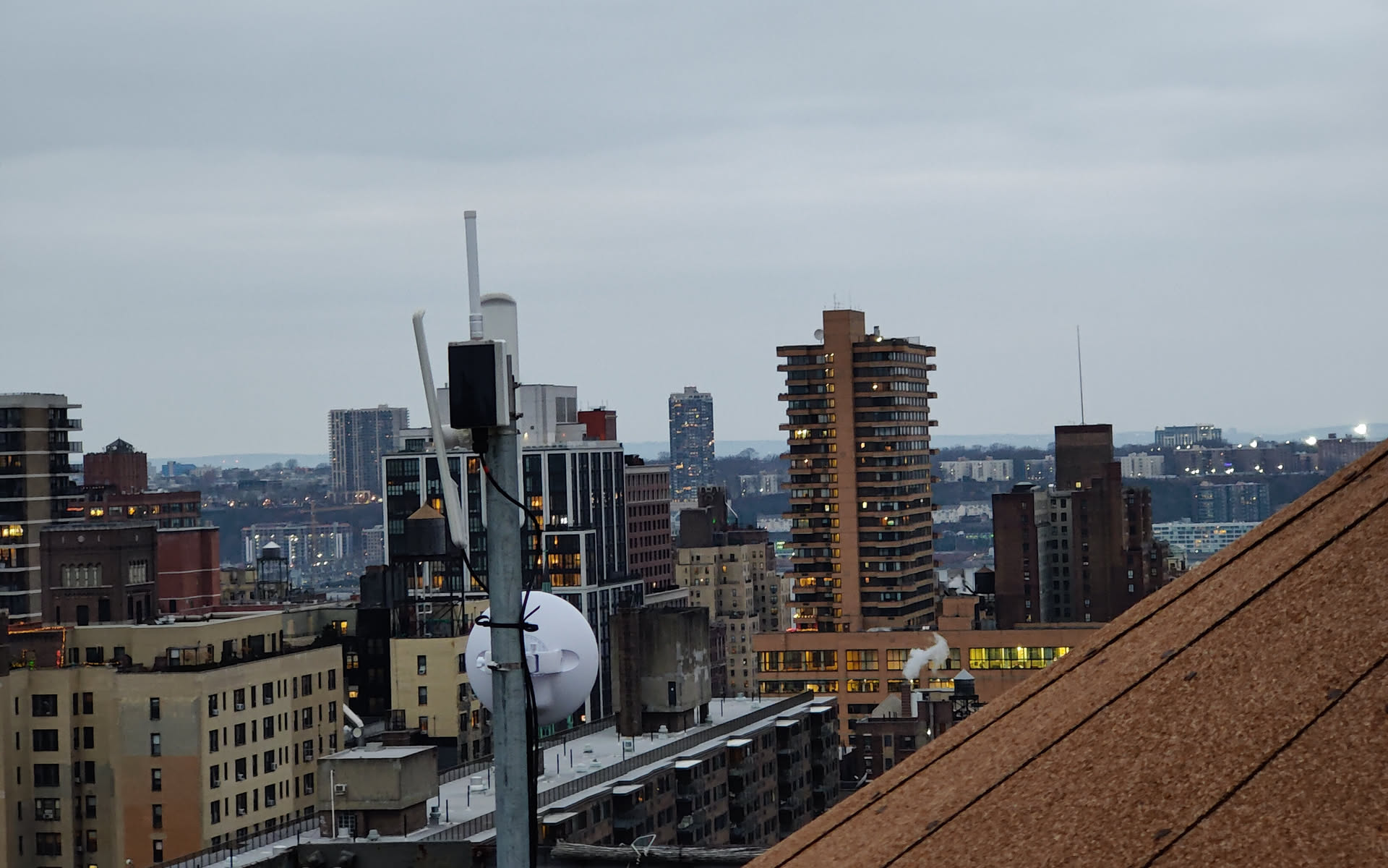 Close-up of a rooftop antenna mast on a NYC Mesh site: a directional dish, a vertical sector antenna, and a small enclosure mounted on a galvanized steel pole. In the background, apartment towers and water tanks fade into an overcast Manhattan afternoon.