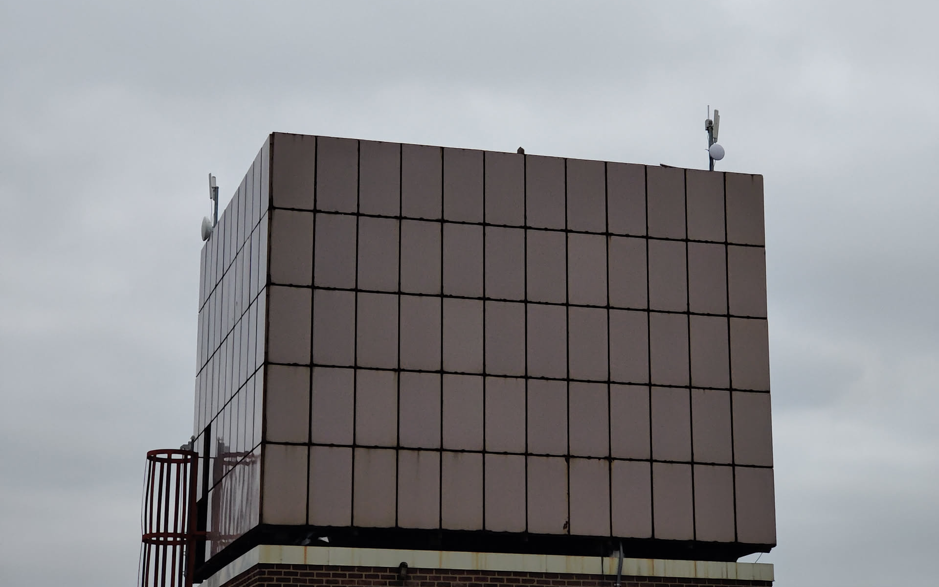A tall paneled rooftop bulkhead — the kind that anchors a NYC Mesh hub — seen from a few yards away under a heavy overcast sky. Small antennas perch on the top corners. A red metal cage ladder runs up the left side. The structure looks like a brutalist concrete monument repurposed for wireless.