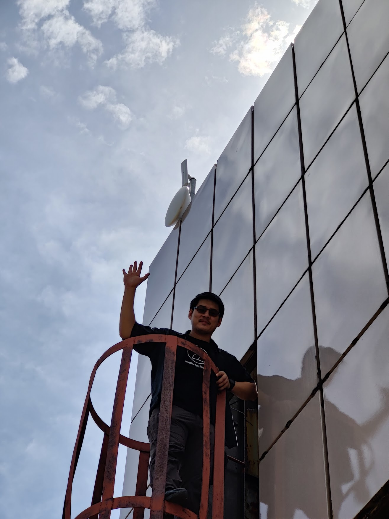 Looking up from below at a tall paneled rooftop bulkhead. A NYC Mesh volunteer in a black 'Audio-Technica' shirt and sunglasses stands on a red metal ladder cage, one arm raised in a wave. A small dish antenna is mounted high on the side of the structure against a partly-cloudy sky.