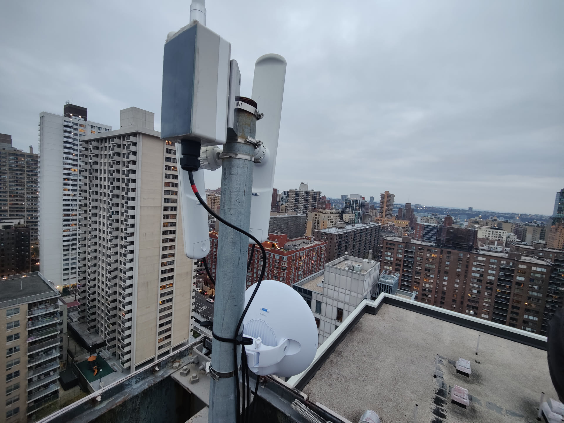 A close-up of another NYC Mesh rooftop antenna cluster: omni-directional cylinder on top, a sector antenna behind, a smaller Wave radio below, all mounted on a steel pole with cables cascading down. In the background, a sea of pre-war Manhattan apartment blocks and water tanks.