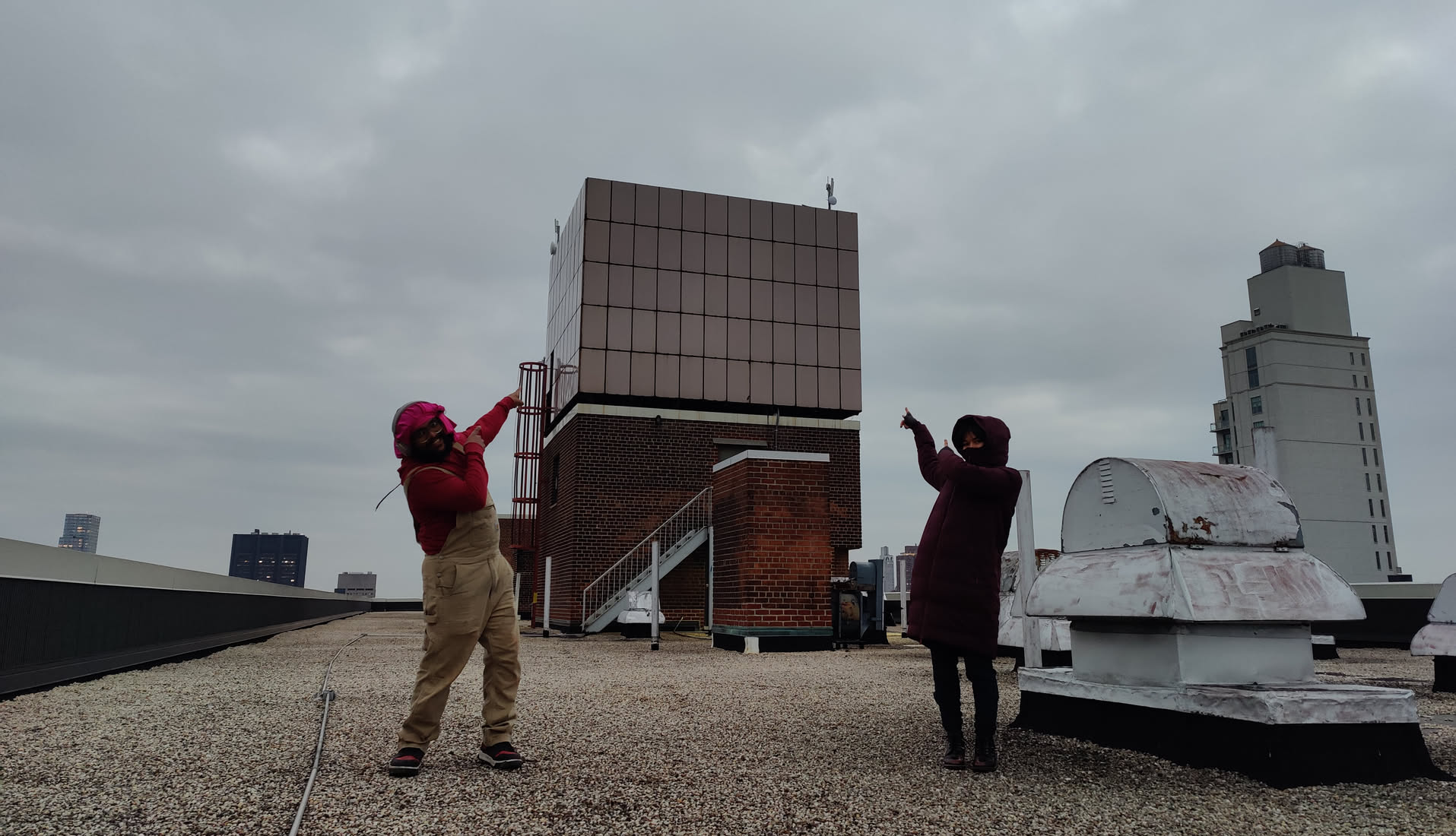 Two NYC Mesh volunteers strike a triumphant pose on a Manhattan rooftop, arms raised, pointing skyward toward a paneled bulkhead with antennas mounted on its corners. Gravel roof, low parapet, a giant water tank to the right, and the distant Midtown skyline. The mood is celebratory — install complete.
