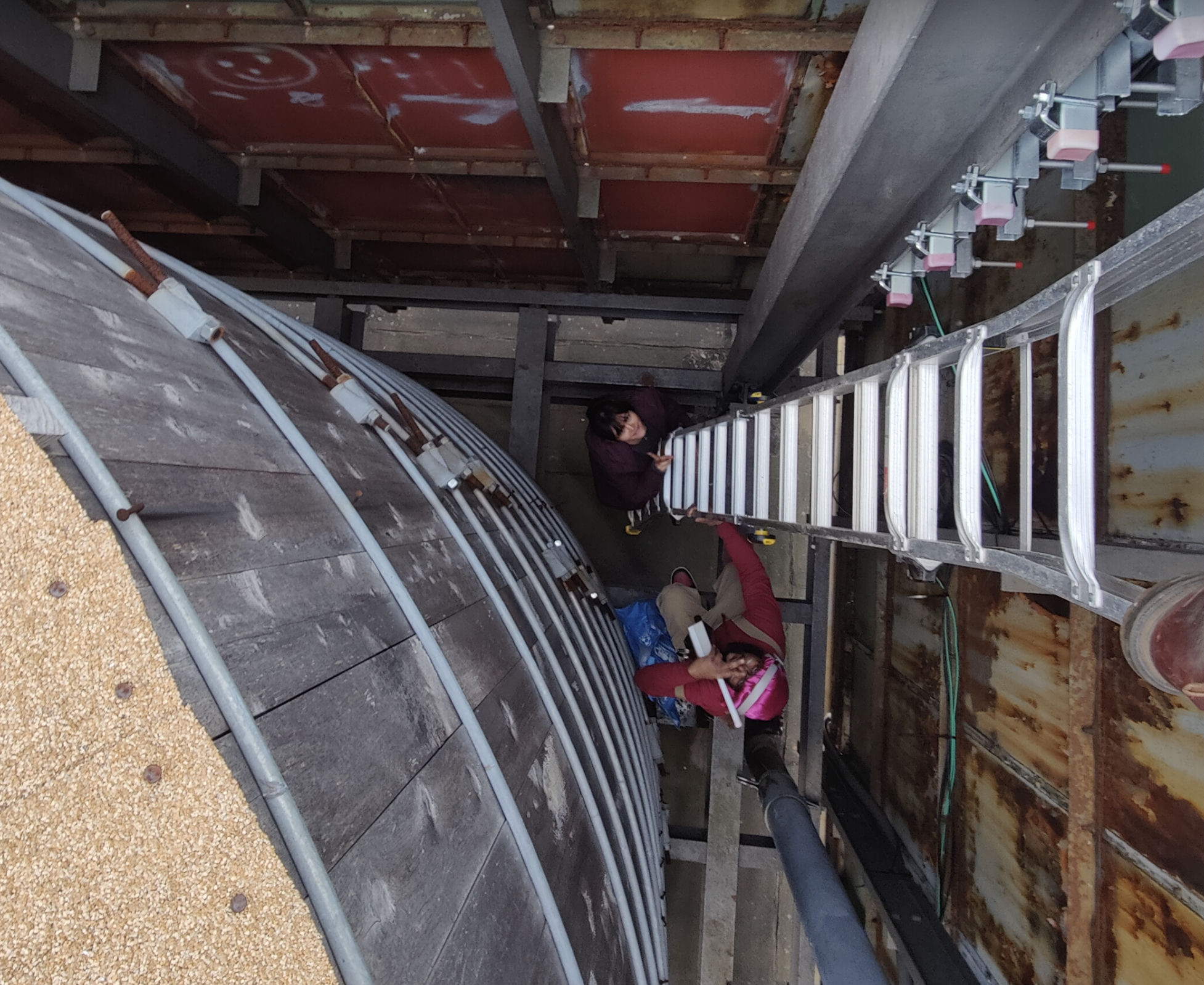 Two volunteers working at the base of a NYC Mesh antenna mount inside a rusted metal-panel rooftop alcove. An aluminum extension ladder cuts diagonally across the frame. One volunteer crouches with tools; the other passes equipment. The scene is workmanlike, intimate, mid-install.