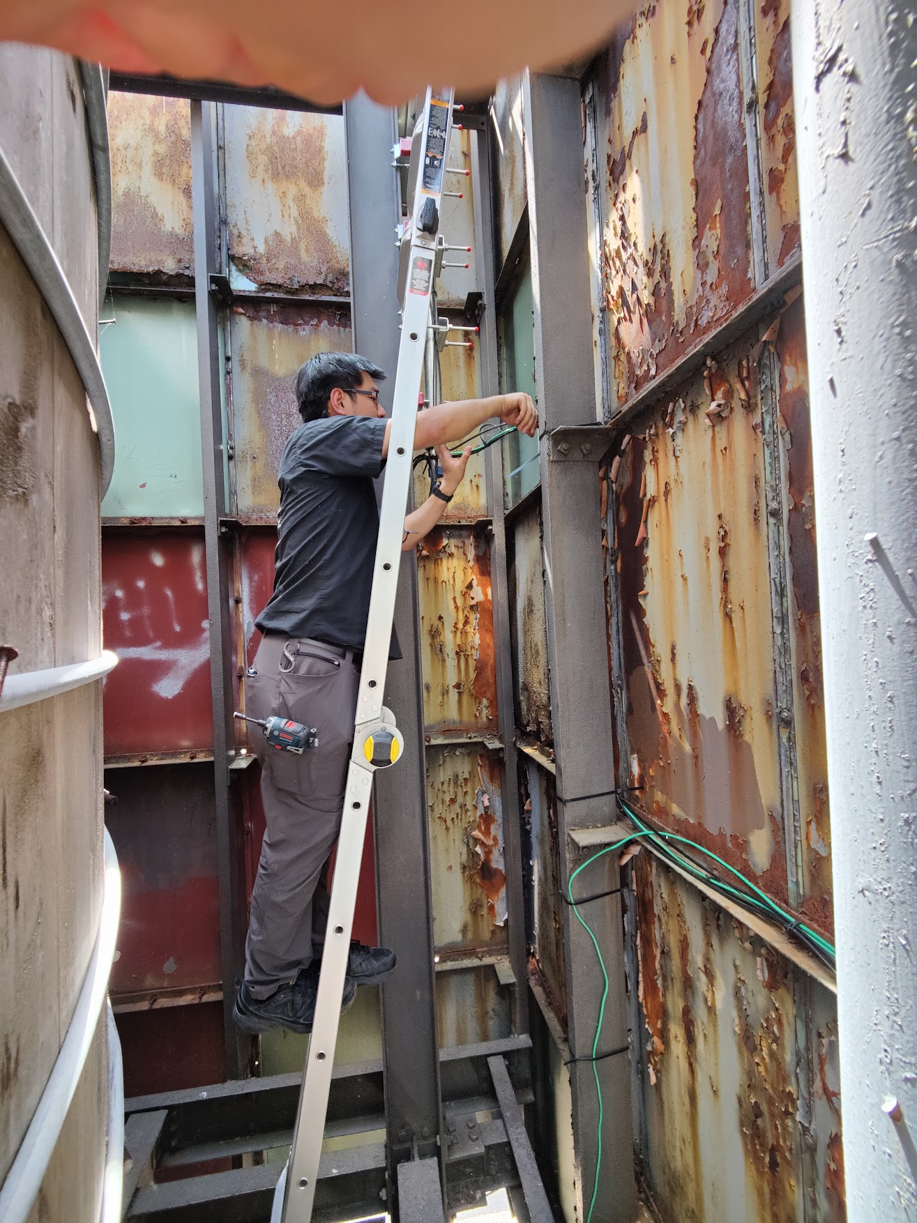 A NYC Mesh volunteer climbs an aluminum extension ladder propped inside a rusted metal-panel bulkhead alcove, a Bosch drill clipped to his belt, working on the cabling and antenna mount above. The scene is gritty, blue-collar, and very real.