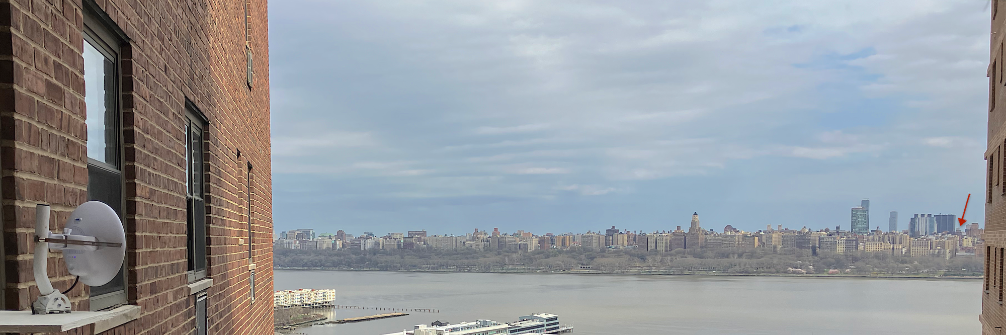 View from a New Jersey apartment window across the Hudson to the Upper West Side skyline. A small white Ubiquiti dish is mounted in the foreground; a red arrow on the right points to a barely-visible building 1.9 miles away on 97th Street.